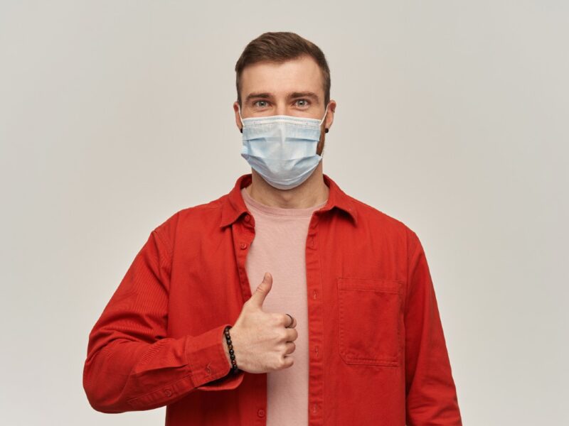confident-young-bearded-man-red-shirt-virus-protective-mask-face-against-coronavirus-standing-showing-thumbs-up-white-wall